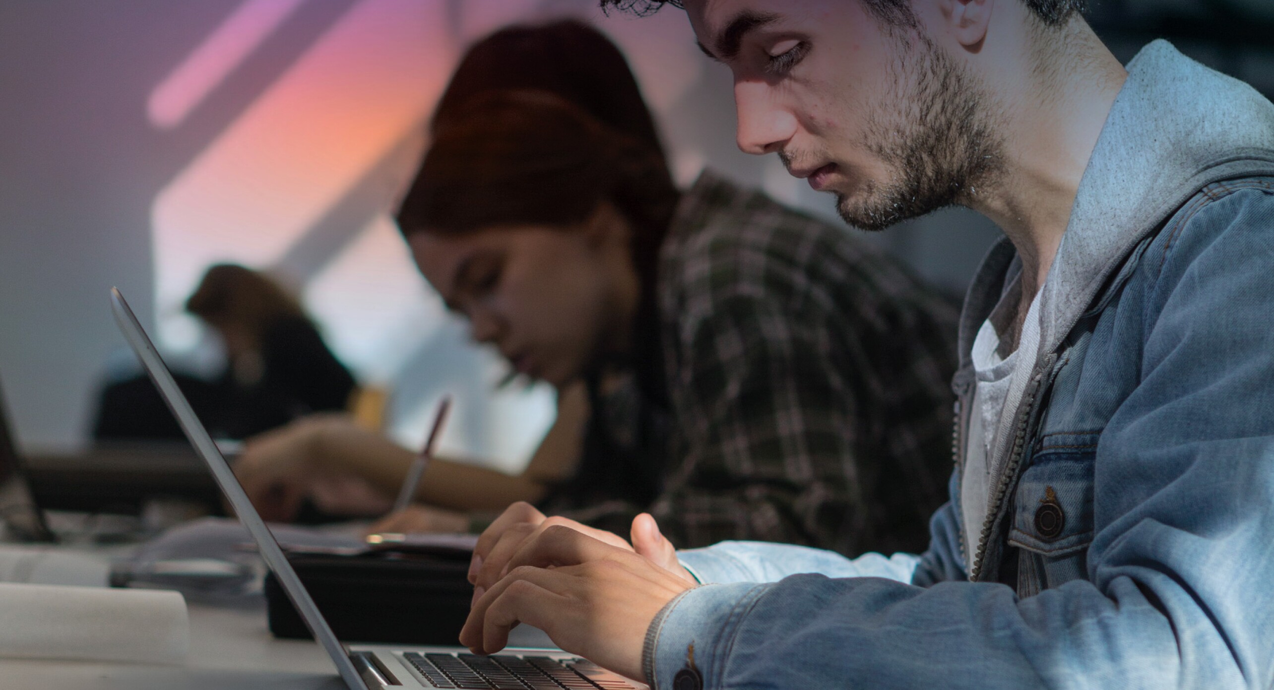 perfil de um rapaz a escrever em computador a uma mesa, com pessoas em fundo a escrever